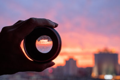 Hand holding camera lens, looking at scenics of glowing clouds at sunset