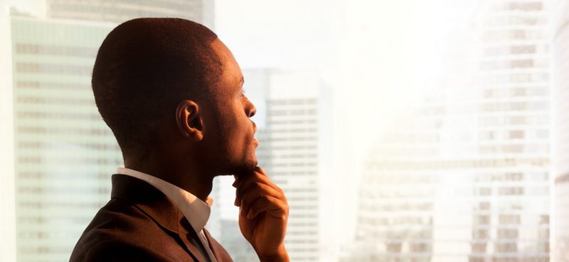 Horizontal photo close up of pensive African American businessman in suit looking out a window big city modern architecture thinking planning future projects.
