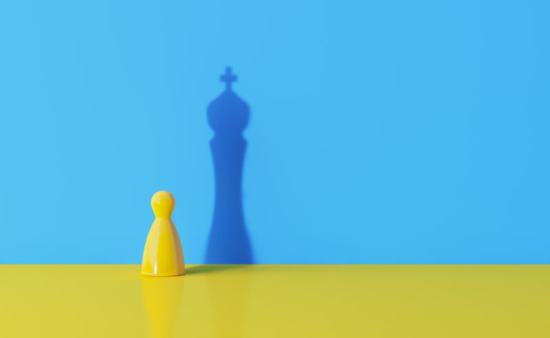 Yellow chess pawn casting the shadow of a king over blue background for a leadership concept.